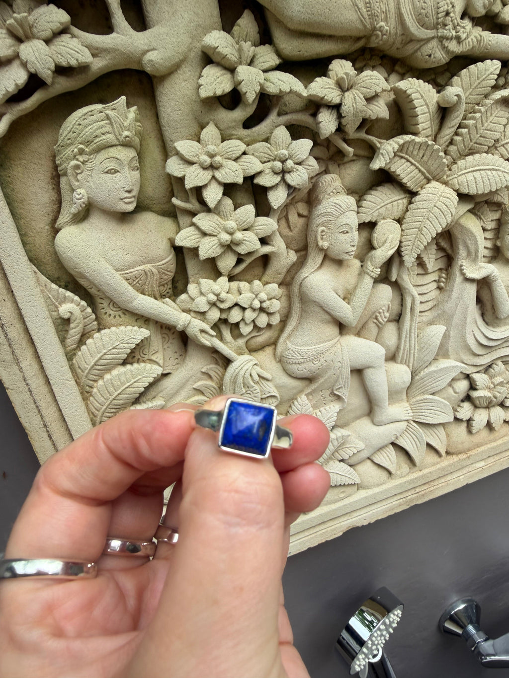 Hand holding a blue stone ring in front of a detailed stone carving.